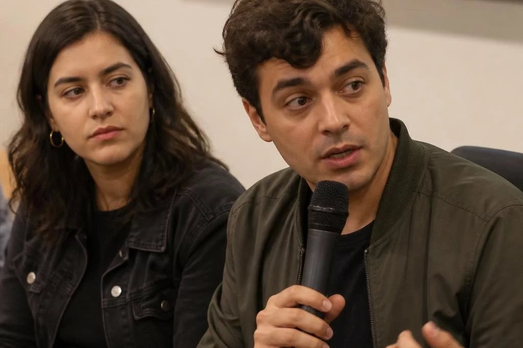 Três participantes conversam ao redor de uma mesa comprida numa sala clara de janelas altas